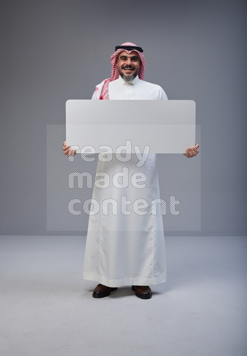 Saudi man Wearing Thob and red Shomag standing holding board on Gray background