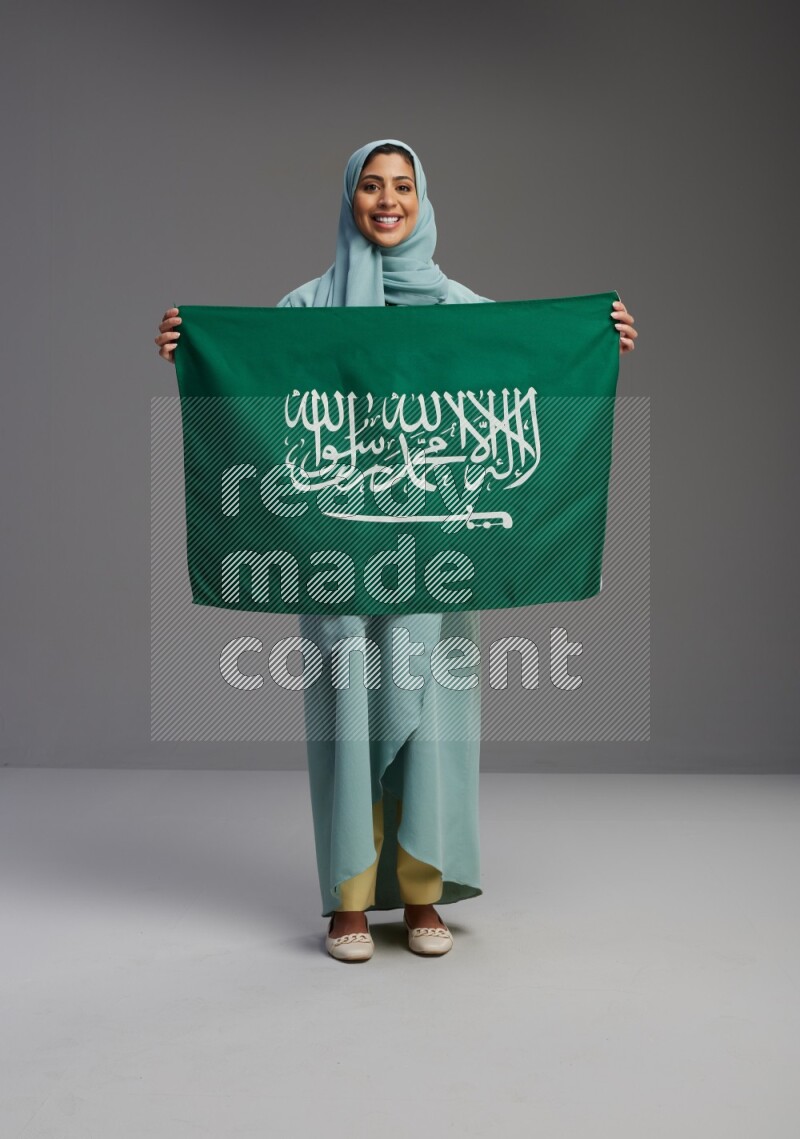 Saudi Woman wearing Abaya standing holding Saudi flag on Gray background