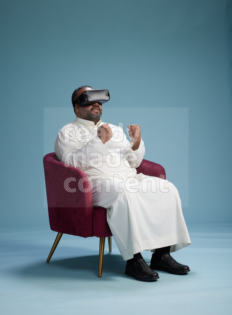 Saudi Man without shimag sitting on chair  gaming with VR on blue background