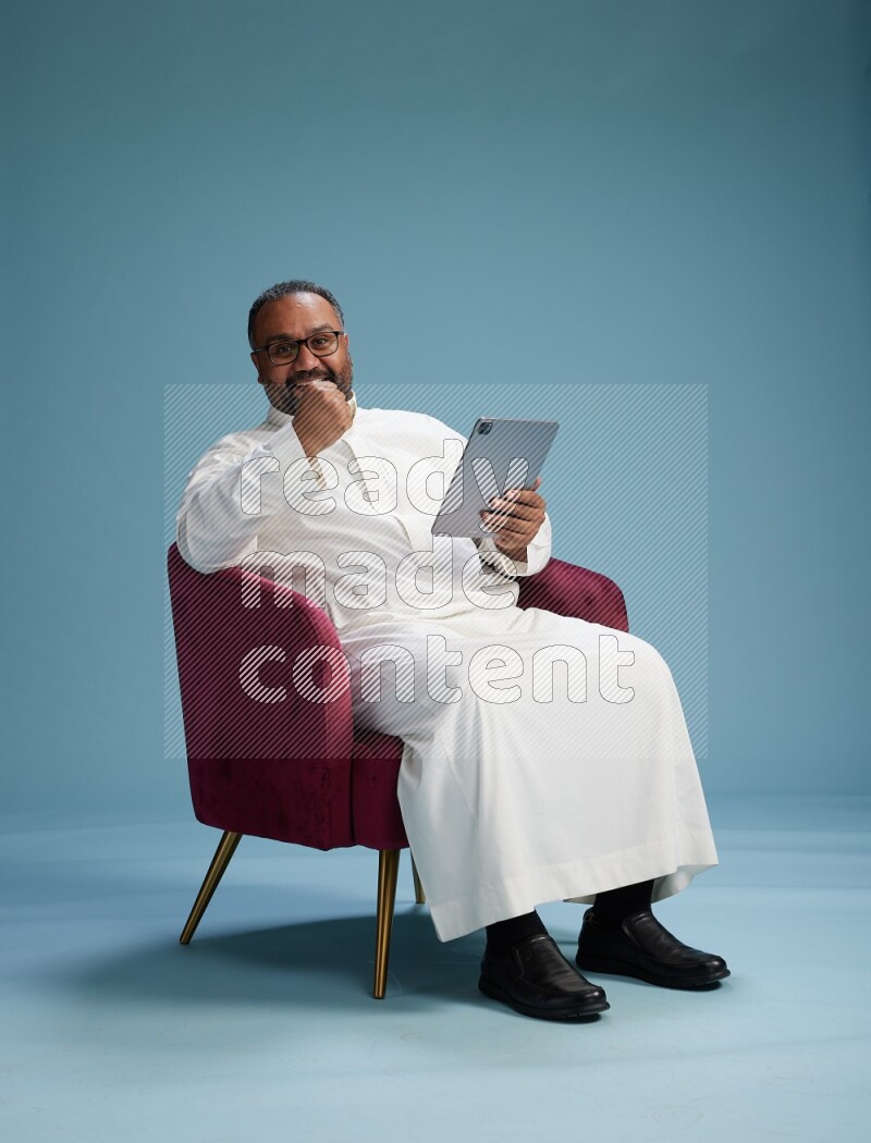 Saudi Man without shimag sitting on chair working on tablet on blue background