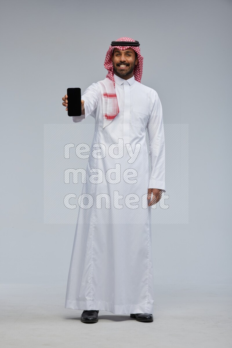 Saudi man Wearing Thob and shomag standing showing phone to camera on Gray background