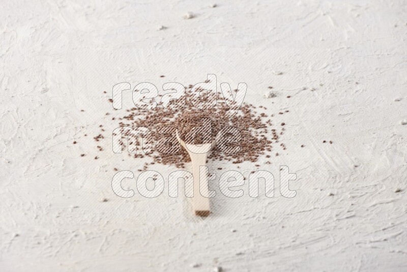 A wooden spoon full of flax seeds surrounded by flax seeds on a textured white flooring