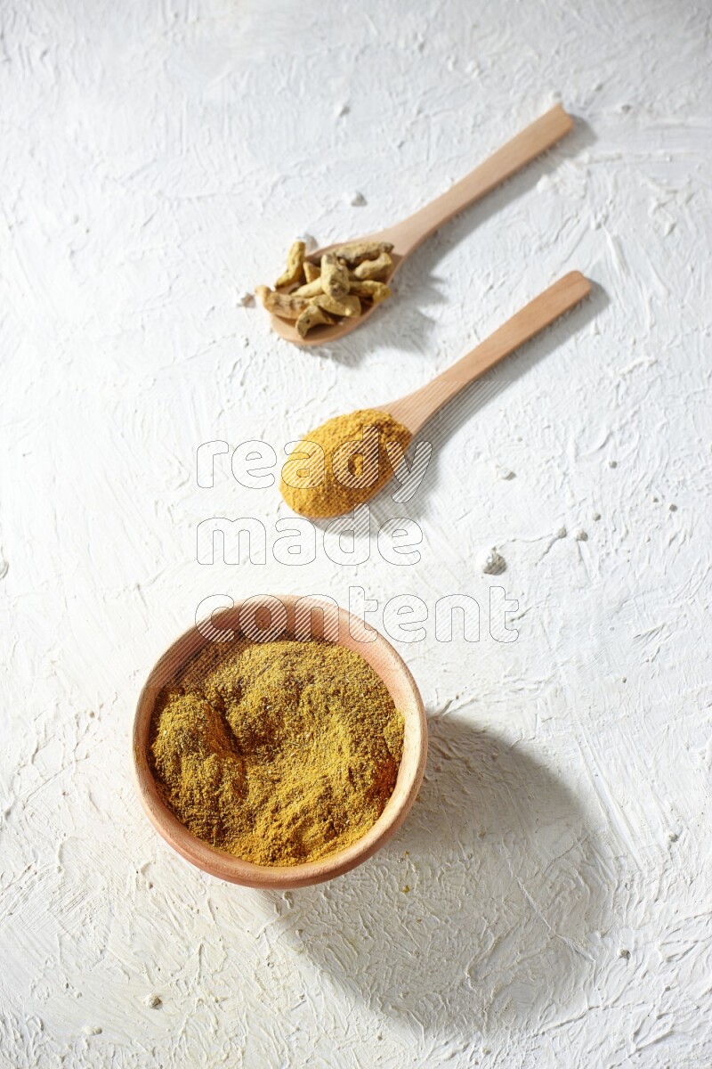 A wooden bowl full of turmeric powder and 2 wooden spoons full of dried turmeric whole finger and turmeric powder on textured white flooring