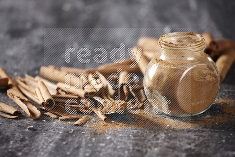 Herbal glass jar and a metal spoon full of cinnamon powder surrounded by cinnamon sticks on textured black background