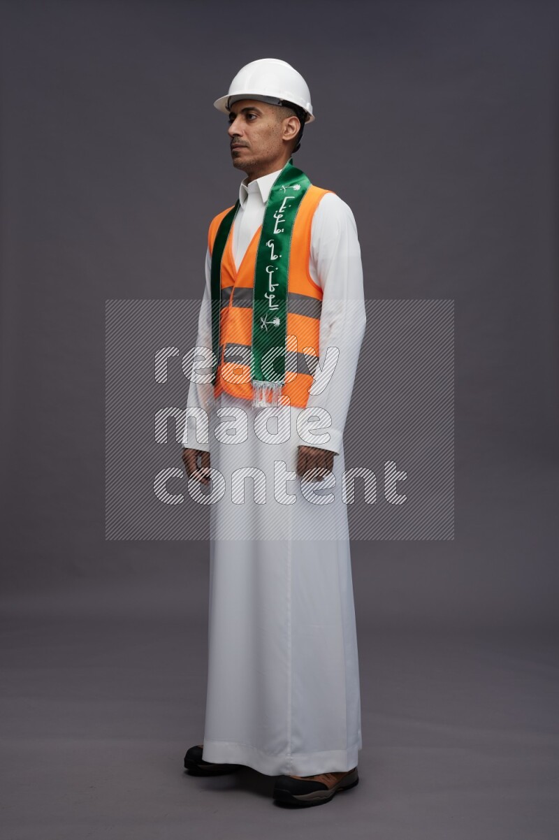 Saudi man wearing thob with engineer vest standing holding Saudi flag on gray background