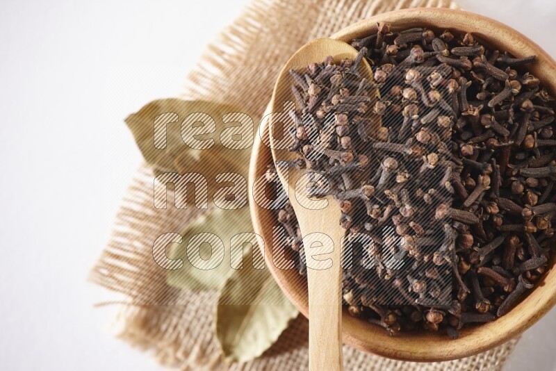 A wooden bowl and a wooden spoon full of cloves on a piece of burlap on a white flooring