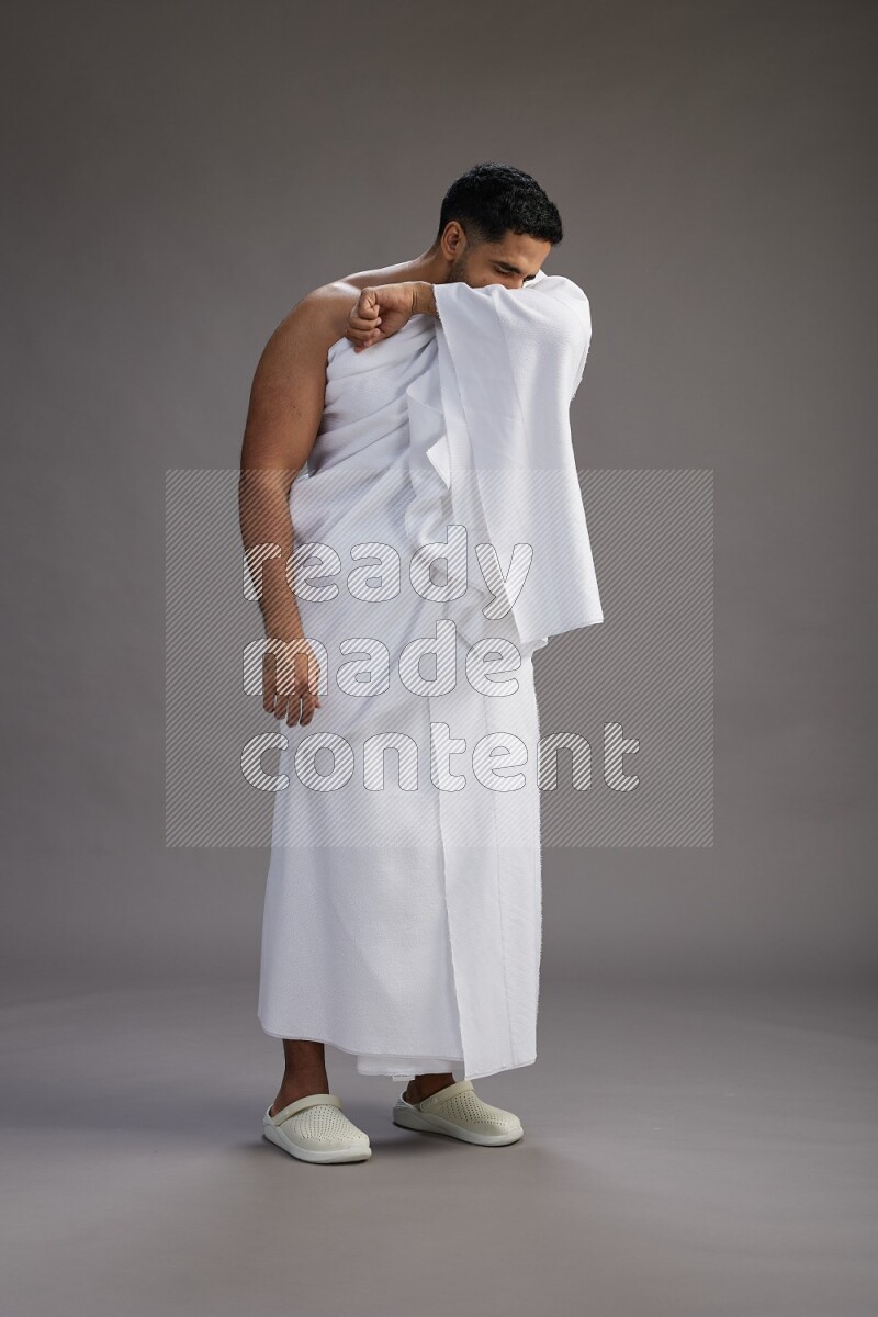 A man wearing Ehram standing showing how to sneeze on gray background