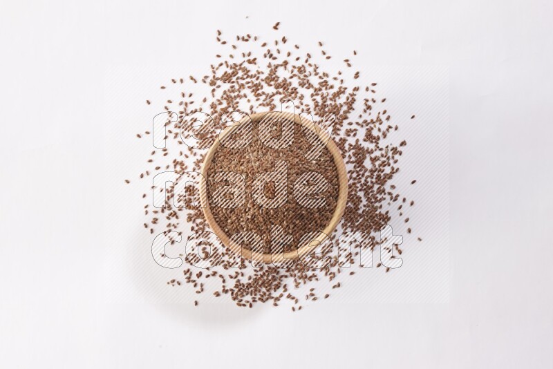 A wooden bowl full of flax seeds surrounded by flax seeds on a white flooring