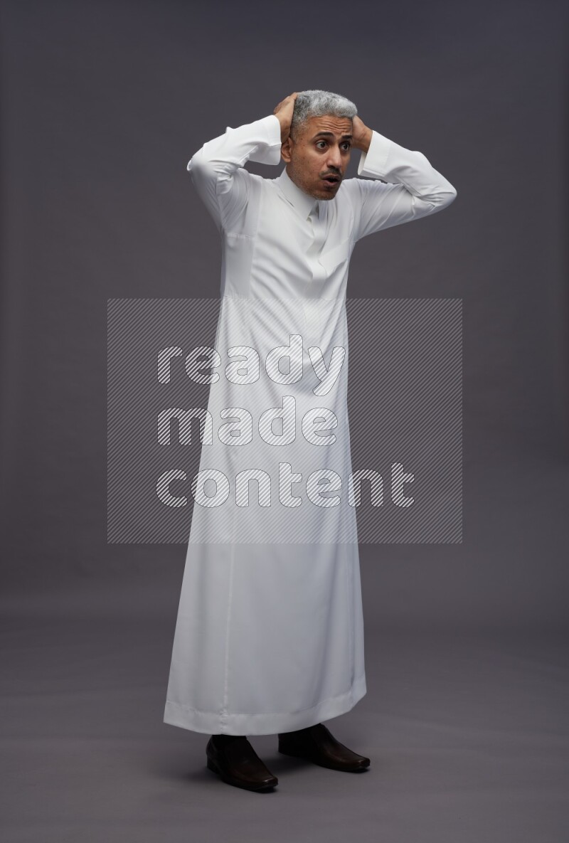 Saudi man wearing thob standing hands behind head on gray background