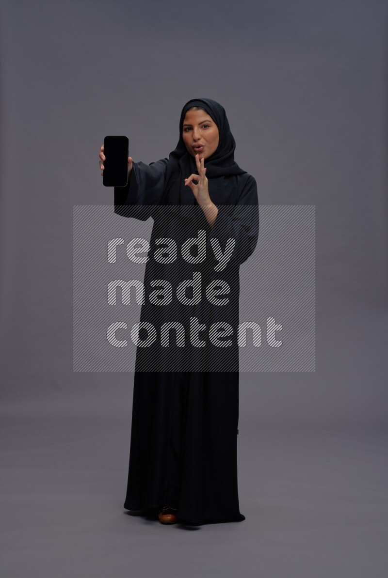 Saudi woman wearing Abaya standing showing phone to camera on gray background
