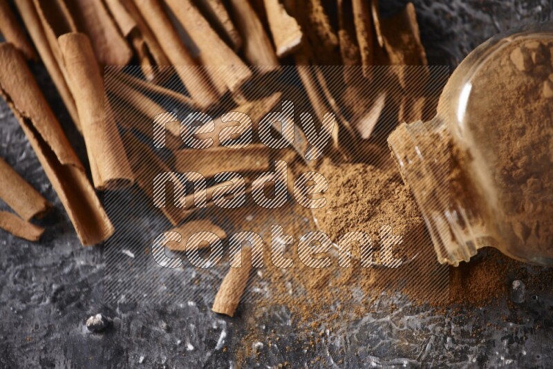 Herbal glass jar full cinnamon powder flipped and a metal spoon full of powder surrounded by cinnamon sticks on textured black background in different angles