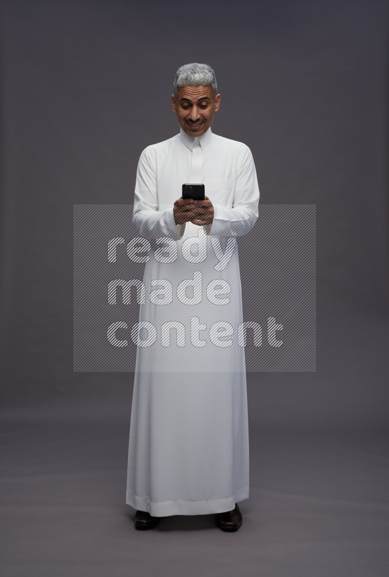 Saudi man wearing thob standing texting on phone on gray background