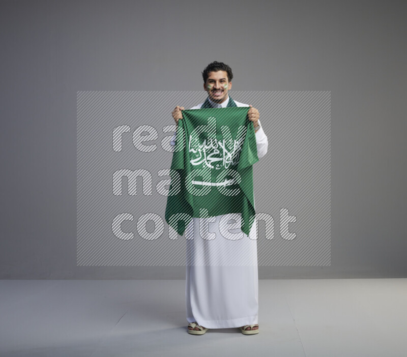 A Saudi man standing wearing thob with face painting wrapping Saudi flag scarf and holding big Saudi flag on gray background