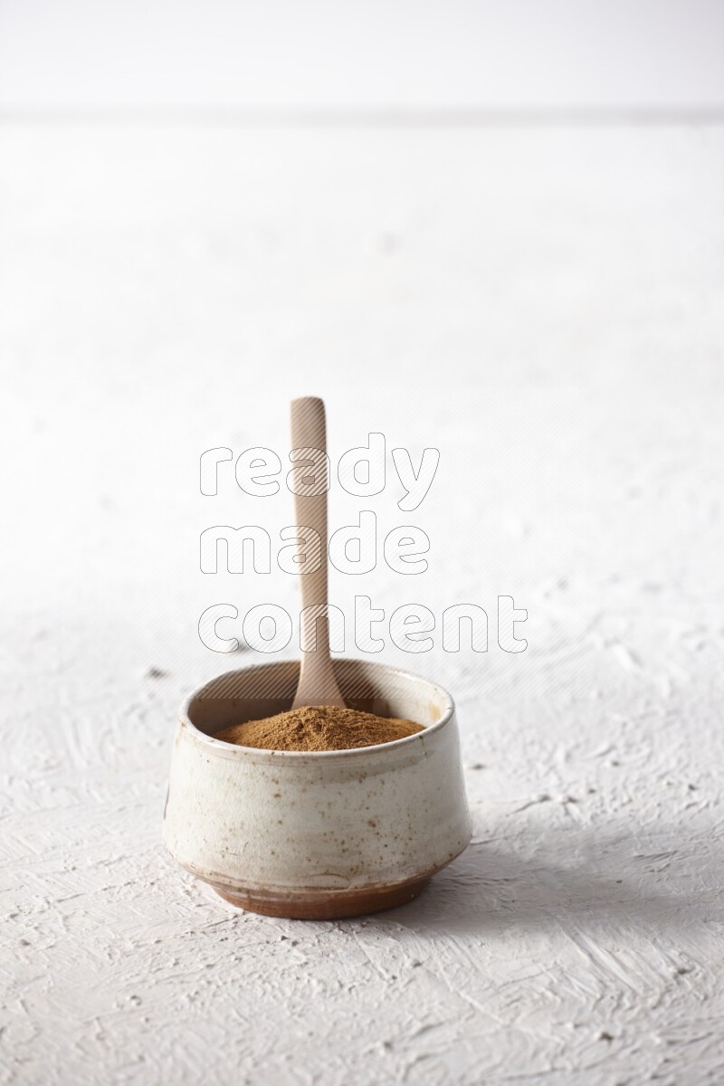 Ceramic beige bowl full of cinnamon powder with a wooden spoon on a textured white background