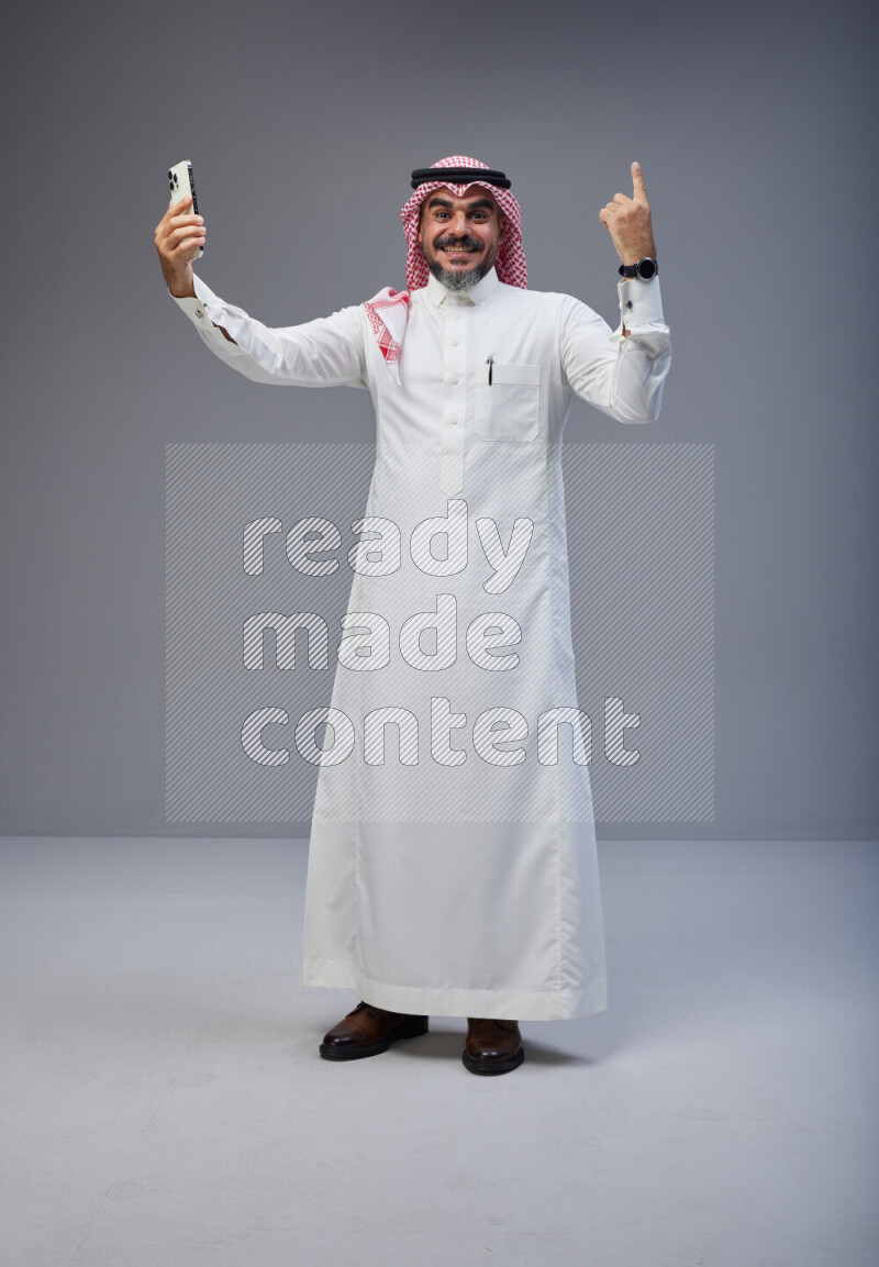Saudi man Wearing Thob and red Shomag standing taking selfie on Gray background