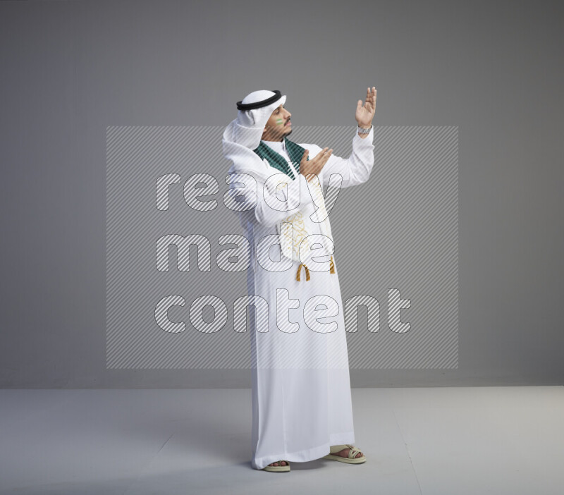 A Saudi man standing wearing thob and white shomag with face painting and Saudi flag scarf on gray background