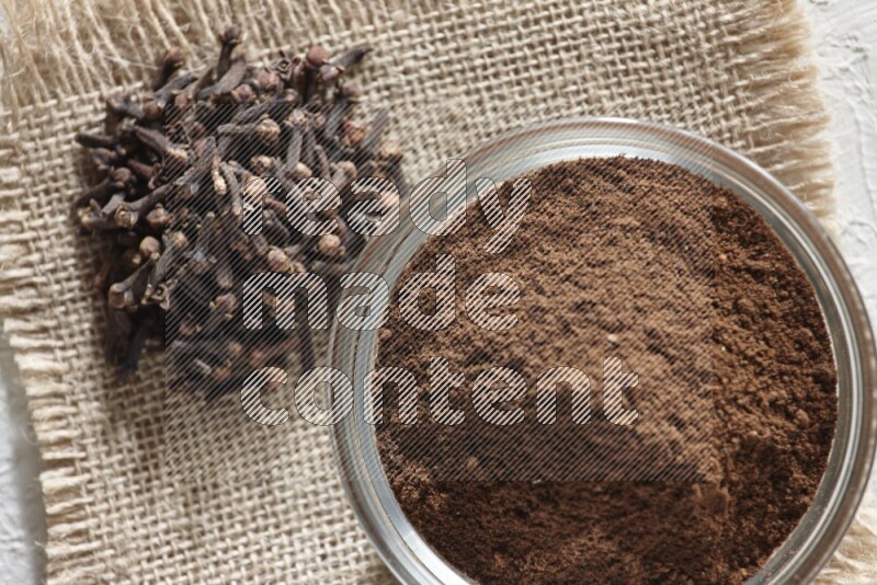 A glass bowl full of cloves powder and cloves grains on a burlap piece on a white flooring