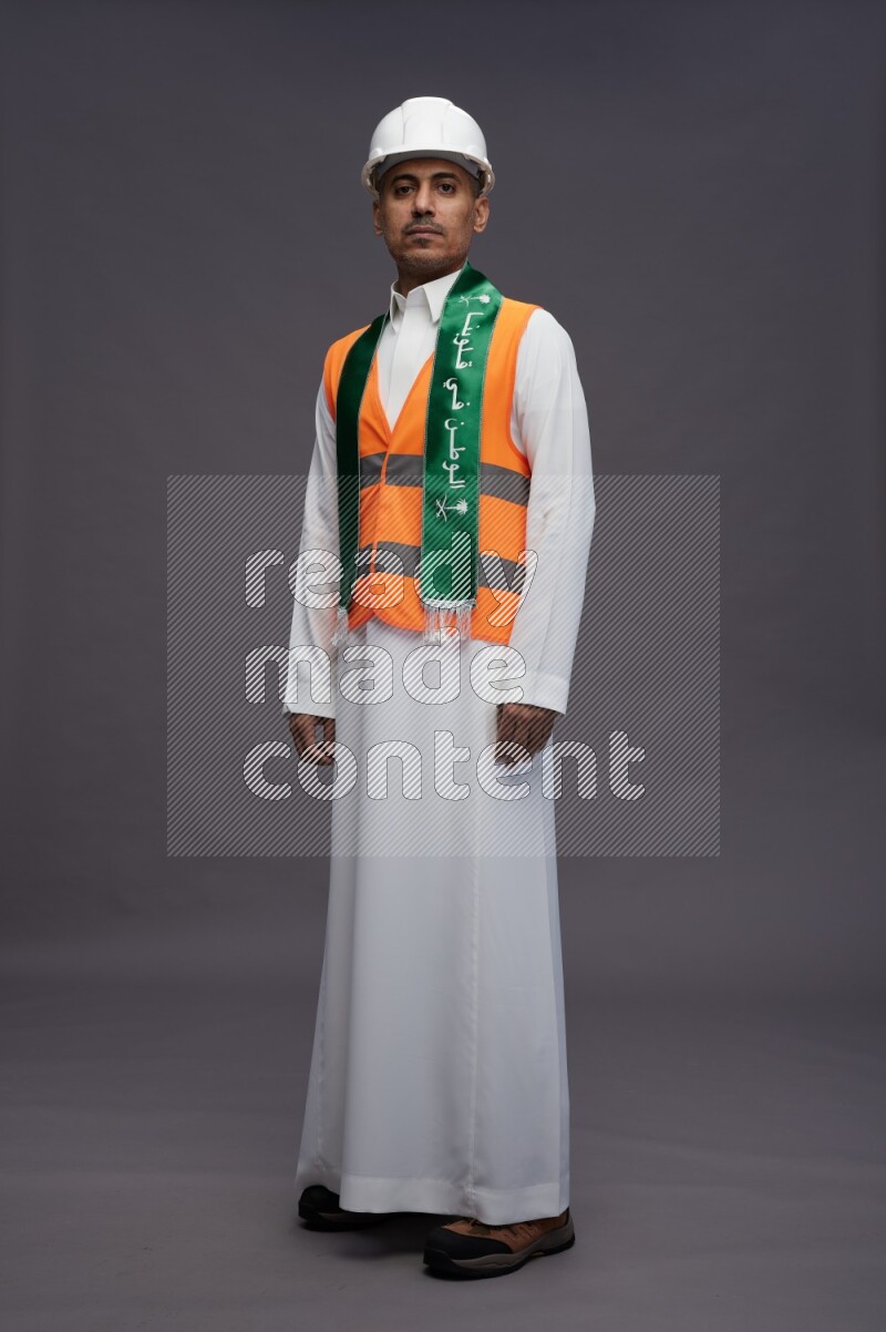 Saudi man wearing thob with engineer vest standing holding Saudi flag on gray background