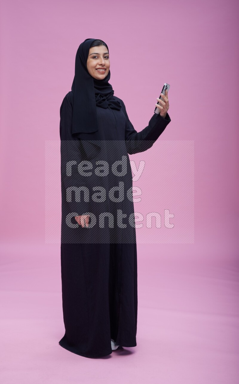 Saudi woman wearing Abaya standing taking selfie on pink background