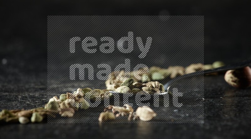 A Metal spoon full of cardamom seeds and some seeds beside it on a textured black flooring