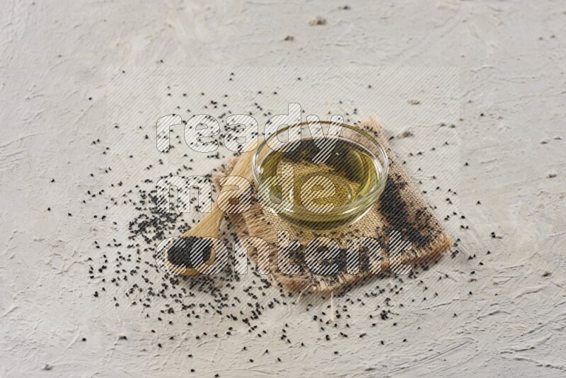A glass bowl full of black seeds oil and wooden spoon full of black seeds with seeds spread on burlap fabric on a textured white flooring