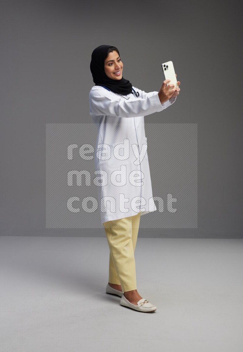 Saudi woman wearing lab coat with stethoscope standing taking selfie on Gray background