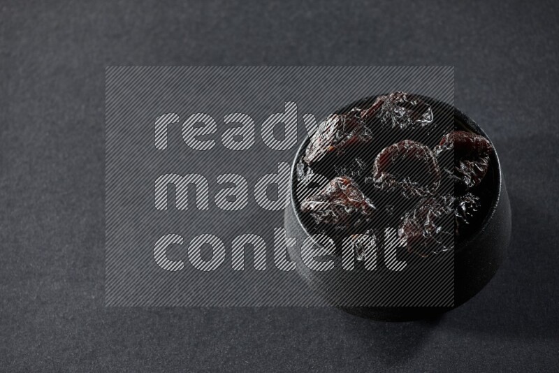 A black pottery bowl full of dried plums on a black background in different angles