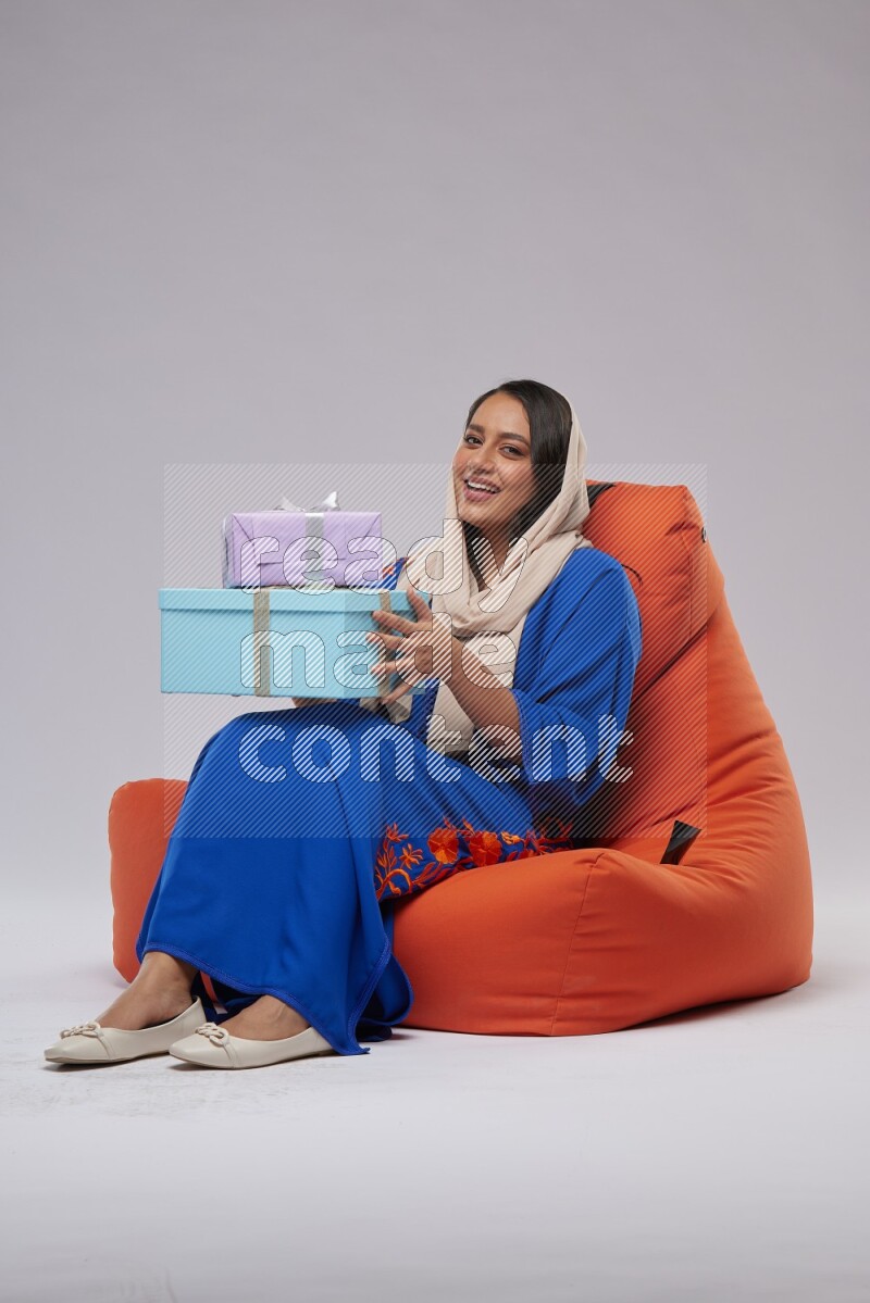 A woman sitting on an orange beanbag wearing Jalabeya holding a gift box