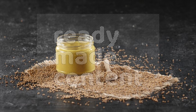 A glass jar full of mustard paste set on a burlap piece with a wooden spoon full of mustard seeds on a textured black flooring