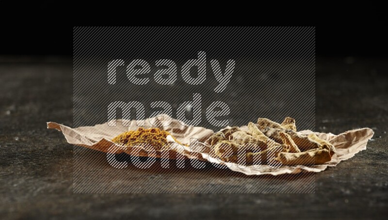 Turmeric powder and dried turmeric whole fingers in 2 crumpled pieces of paper on textured black flooring