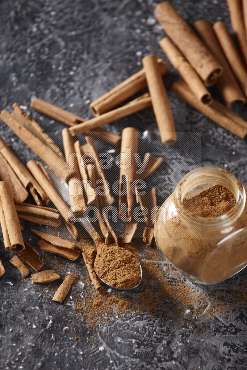 Herbal glass jar and a metal spoon full of cinnamon powder surrounded by cinnamon sticks on textured black background