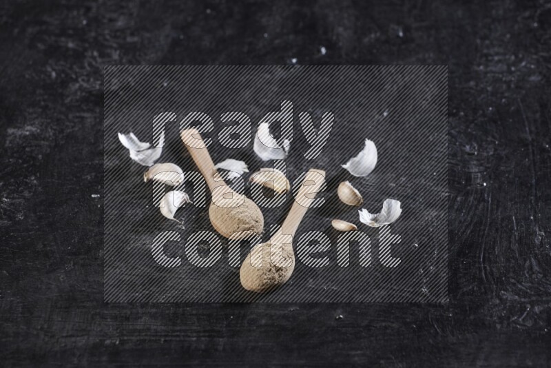 Two wooden spoons filled with garlic powder, surrounded by peeled garlic cloves and their skins on a textured black flooring