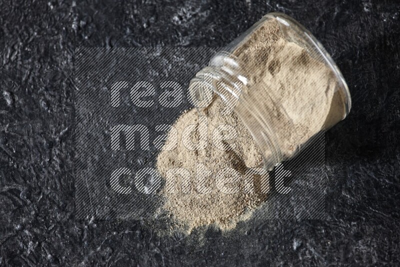 A flipped glass jar full of white pepper powder with spilled powder on textured black flooring