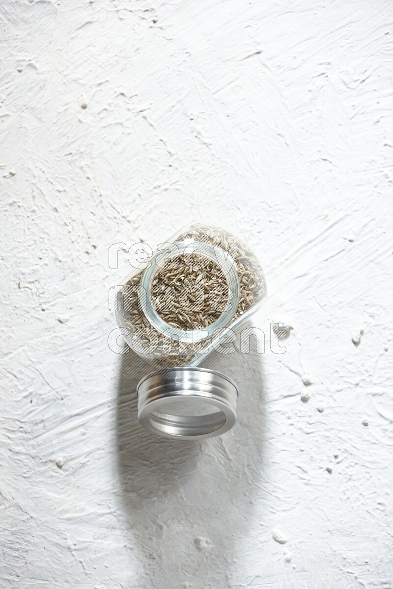 A glass spice jar full of cumin seeds on textured white flooring