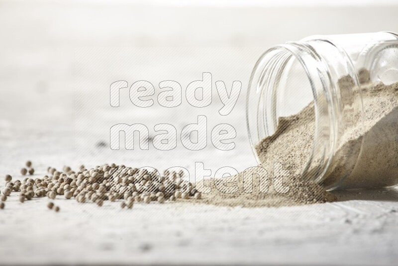 A flipped glass jar full of white pepper powder with spilled powder and white pepper beads on textured white flooring
