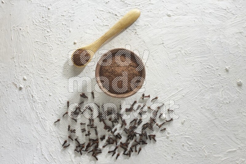 A wooden bowl and wooden spoon full of cloves powder with cloves spread on textured white flooring
