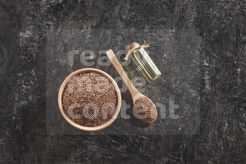 A wooden bowl and spoon full of flaxseeds and a glass bottle of flaxseeds oil on a textured black flooring