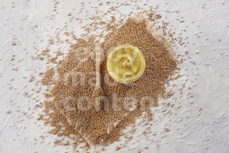 A glass jar full of mustard paste set on a burlap piece with a wooden spoon full of mustard seeds on a textured white flooring