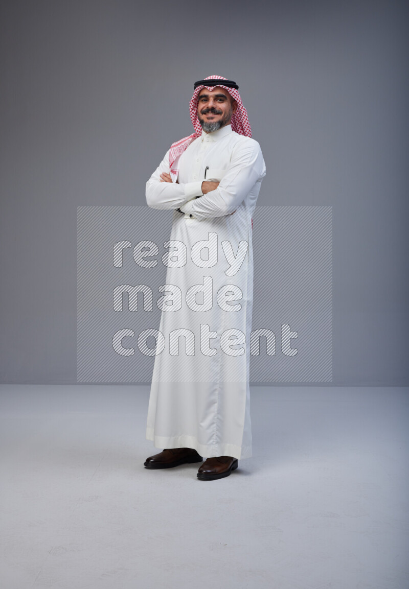 Saudi man Wearing Thob and red Shomag standing with crossed arms on Gray background