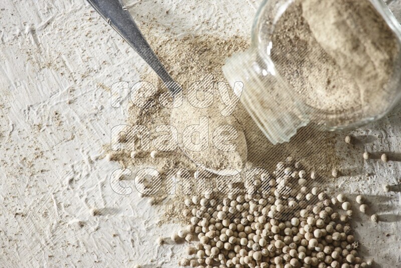 A flipped herbal glass jar and metal spoon full of white pepper powder with spilled powder and beads on textured white flooring