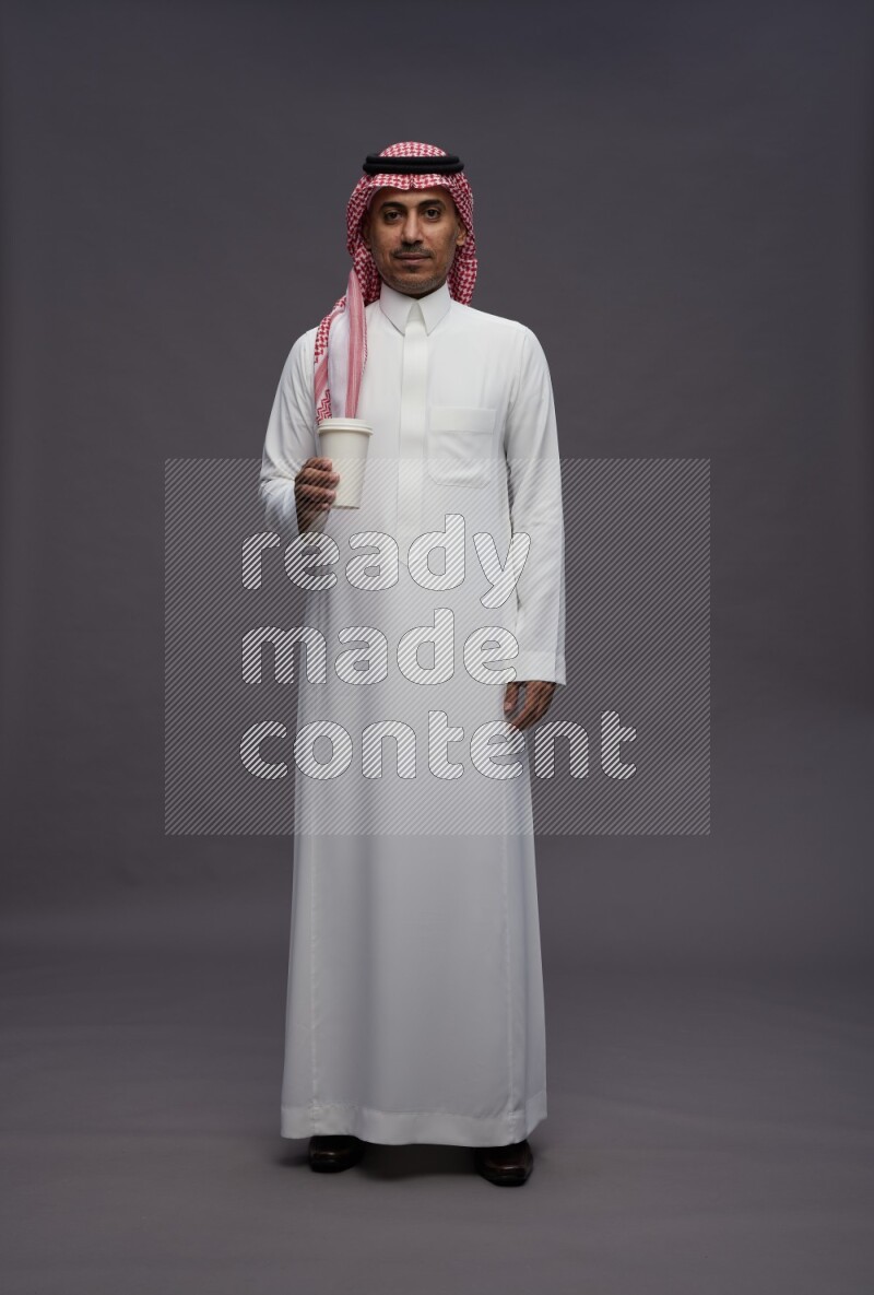 Saudi man wearing thob and shomag standing holding paper cup on gray background