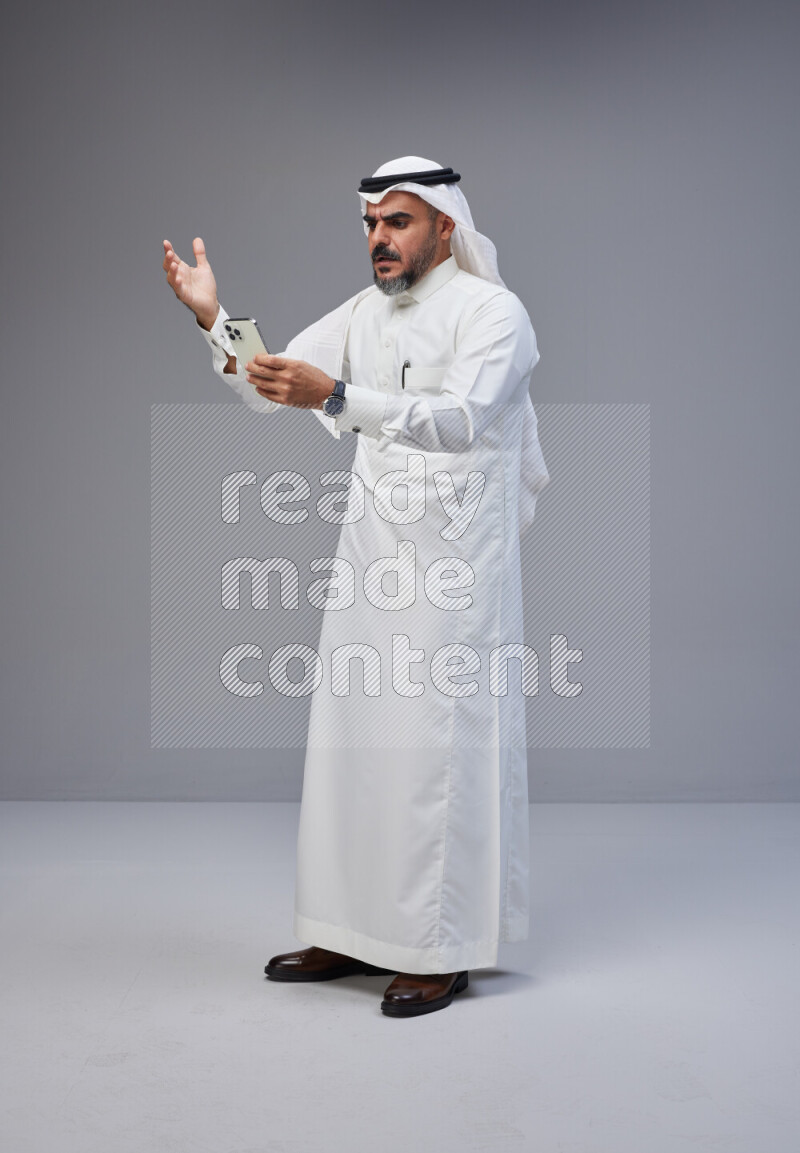 Saudi man Wearing Thob and white Shomag standing texting on phone on Gray background