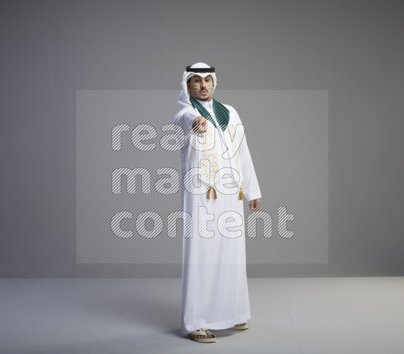 A Saudi man standing wearing thob and white shomag with face painting and Saudi flag scarf on gray background