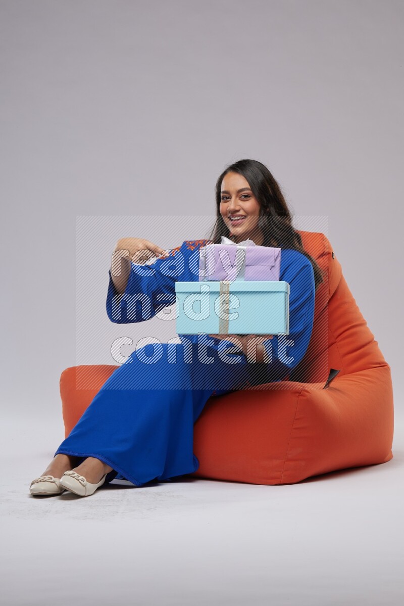 A woman sitting on an orange beanbag wearing Jalabeya holding a gift box
