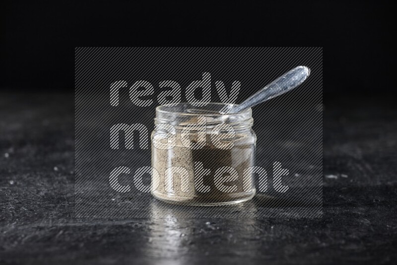 A glass jar and a metal spoon full of white pepper powder on textured black flooring