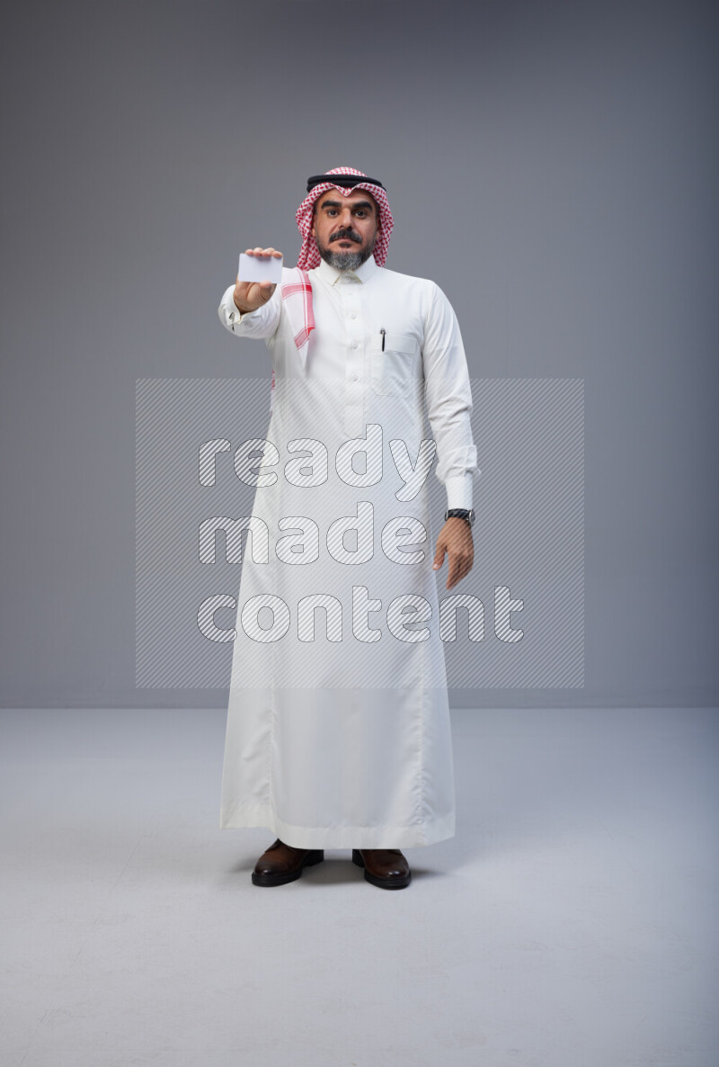 Saudi man Wearing Thob and red Shomag standing holding ATM card on Gray background