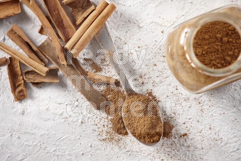 Herbal glass jar full cinnamon powder and a metal spoon surrounded by cinnamon sticks on a white background