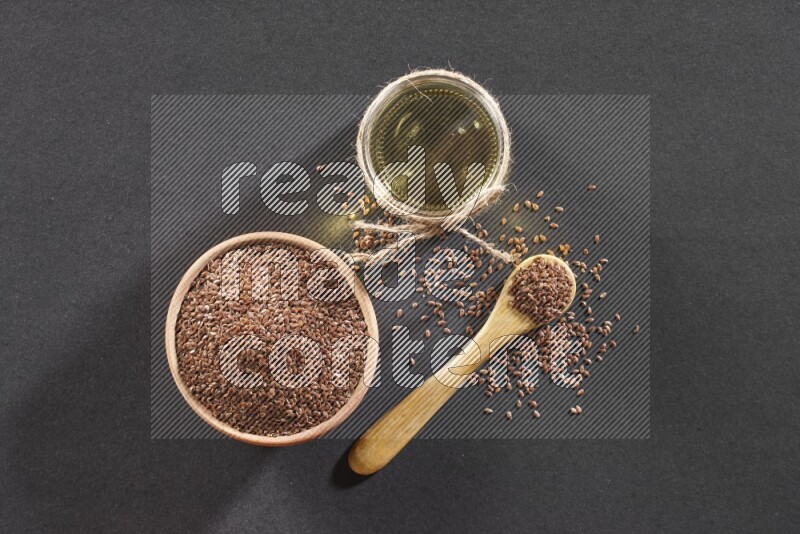 A wooden bowl and wooden spoon full of flaxseeds with a glass jar of flaxseeds oil on a black flooring