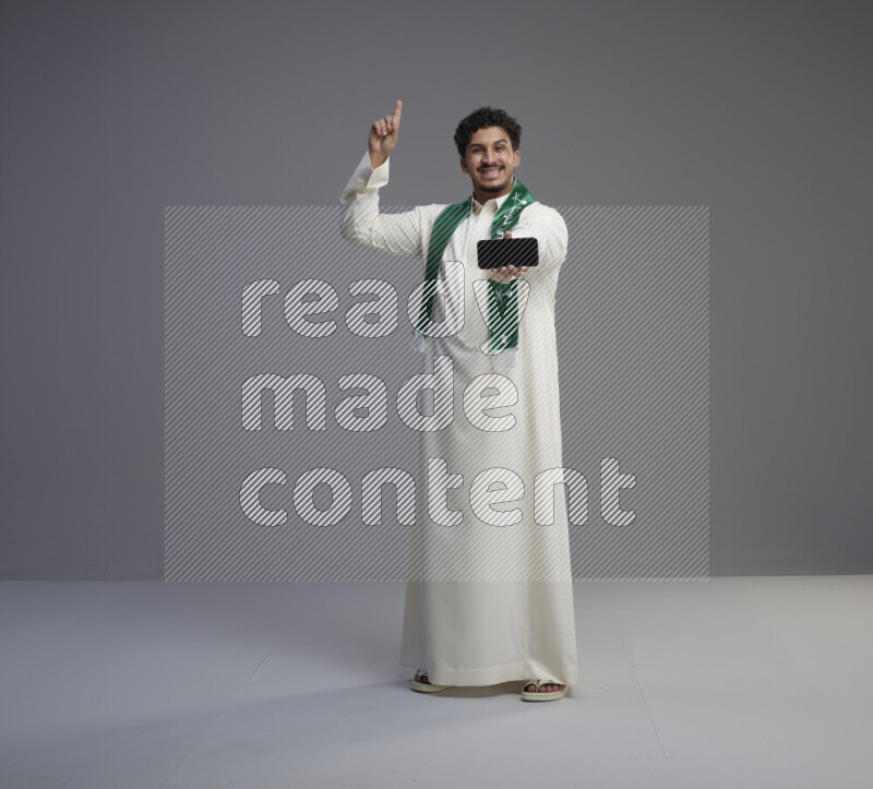 A Saudi man standing wearing thob and Saudi flag scarf showing phone to camera on gray background