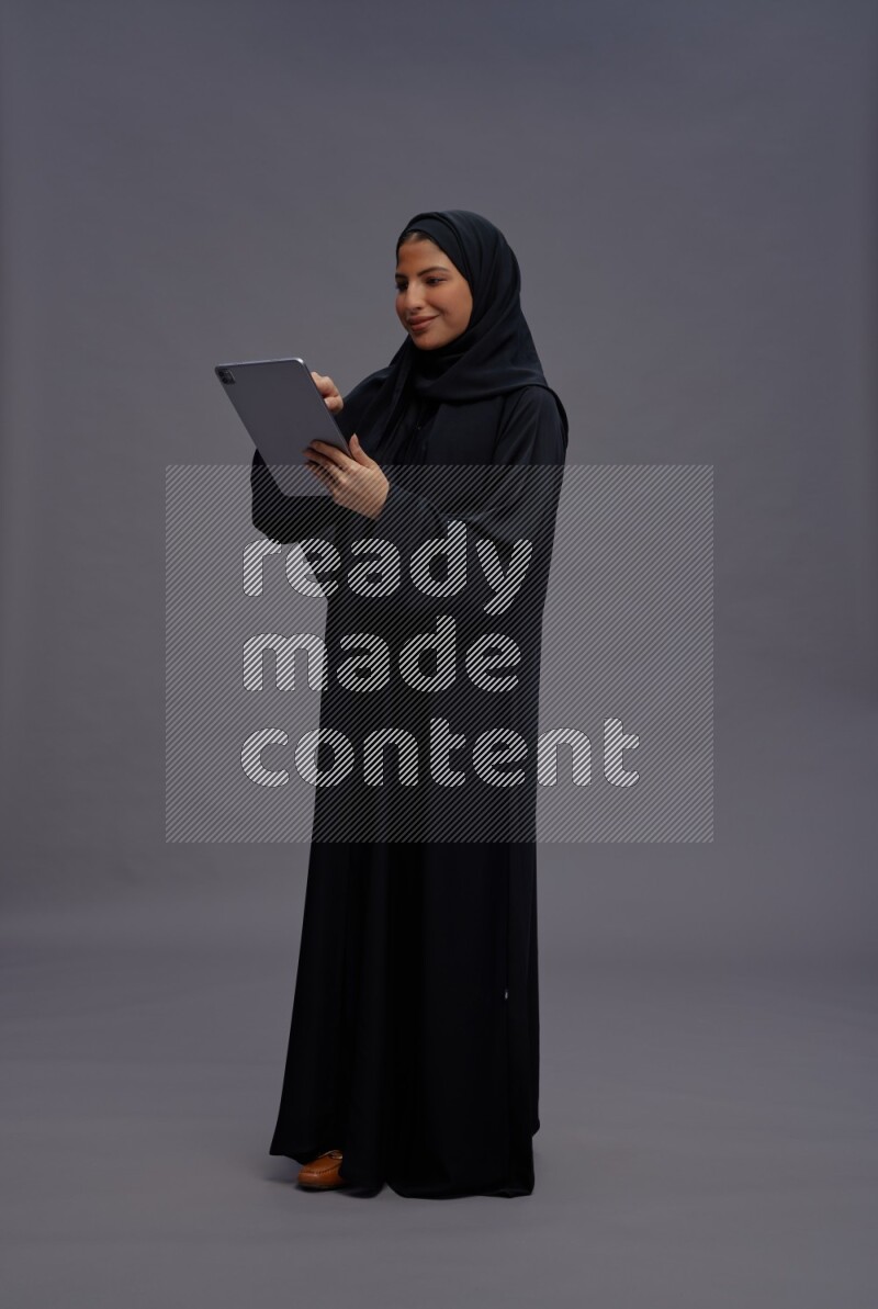 Saudi woman wearing Abaya standing working on tablet on gray background